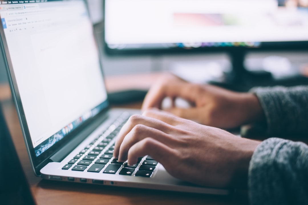 Bir kişinin dizüstü bilgisayar klavyesinde yazdığı ellerin yakın çekimi, arka planda bir masada başka bir bilgisayar monitörü görünür—WordPress Hosting görevlerini yönetmek için mükemmel.